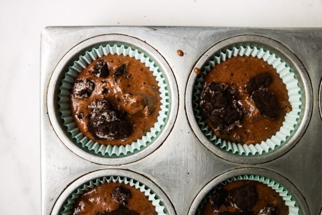 This is an overhead horizontal image of silver muffin tin on a white surface. Chocolate batter is in muffin liners with crushed oreos is in each muffin cup.