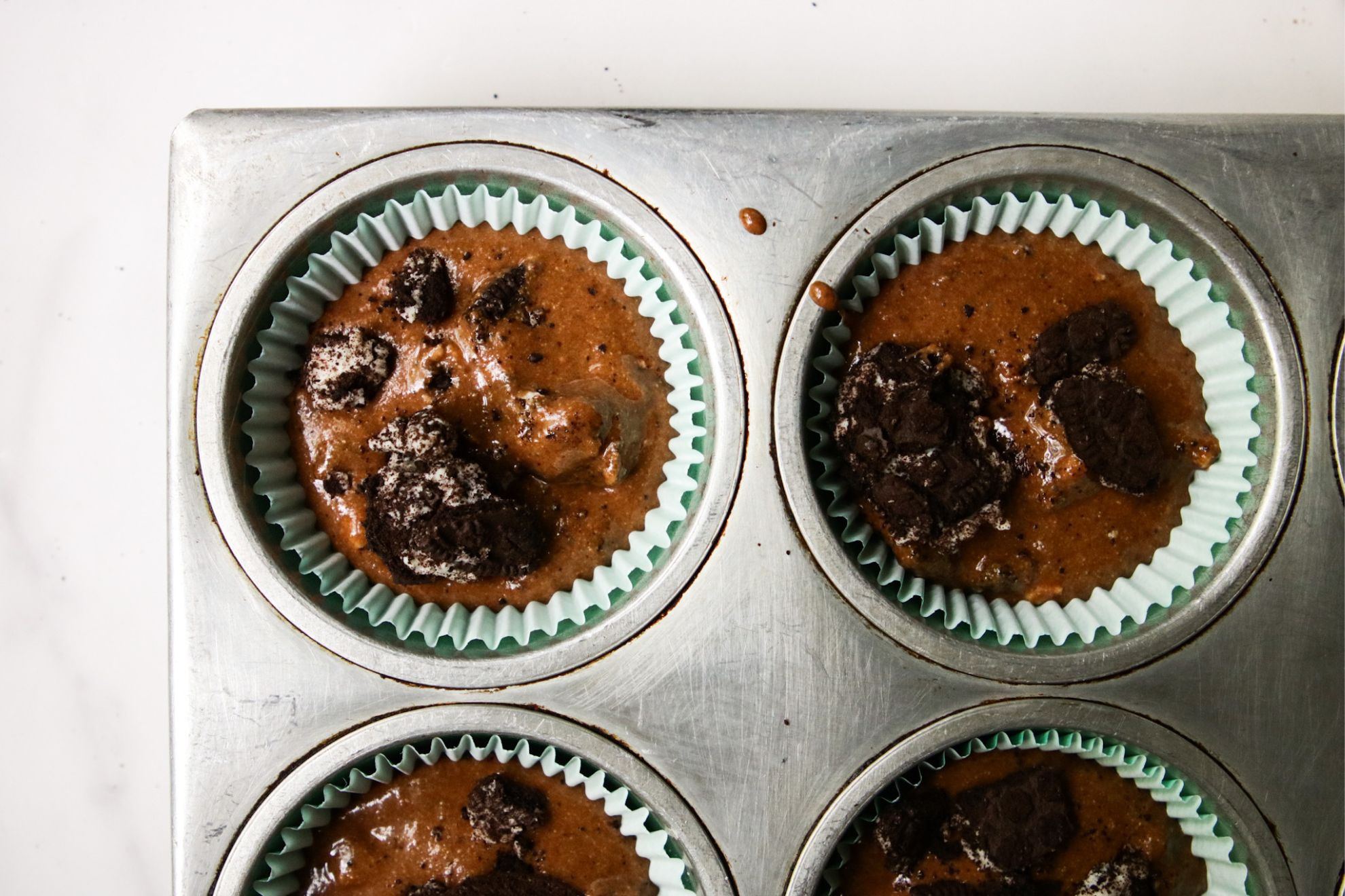 This is an overhead horizontal image of silver muffin tin on a white surface. Chocolate batter is in muffin liners with crushed oreos is in each muffin cup.