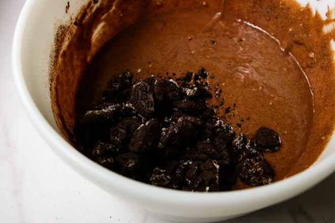 This is a horizontal image of a side view of a large white bowl with chocolate batter and crushed oreos in it. The white bowl sits on a white marble surface.