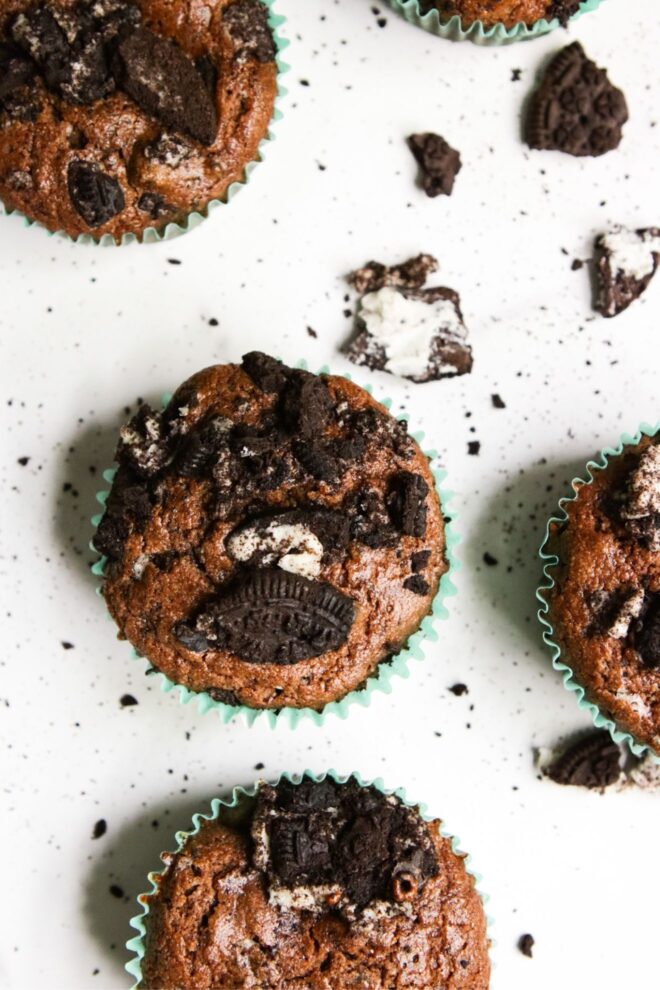 This is an overhead vertical image looking down on the tops of chocolate muffins top with crushed oreos. The muffins are on a white surface with more crushed oreos around them. The muffins have teal liners.