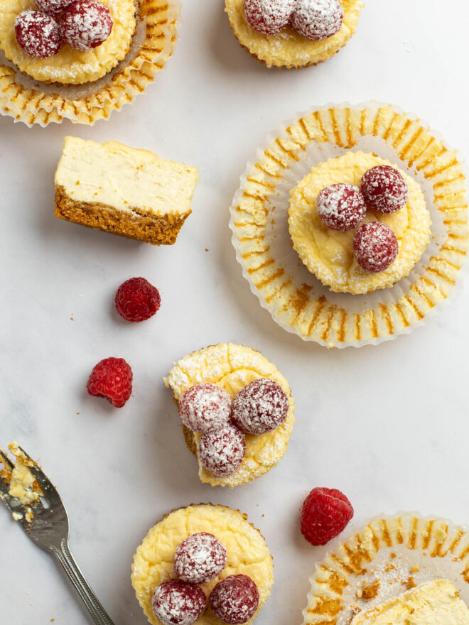 This is an overhead vertical image of mini cheesecakes baked in a muffin tin. Some of the cheesecakes have muffin lined pulled away from the sides. The cheesecakes are topped with raspberries and powdered sugar. One is cut in half and laid on its side to reveal the middle and cookie crust. More raspberries are on the white marble surface around the cheesecakes. 