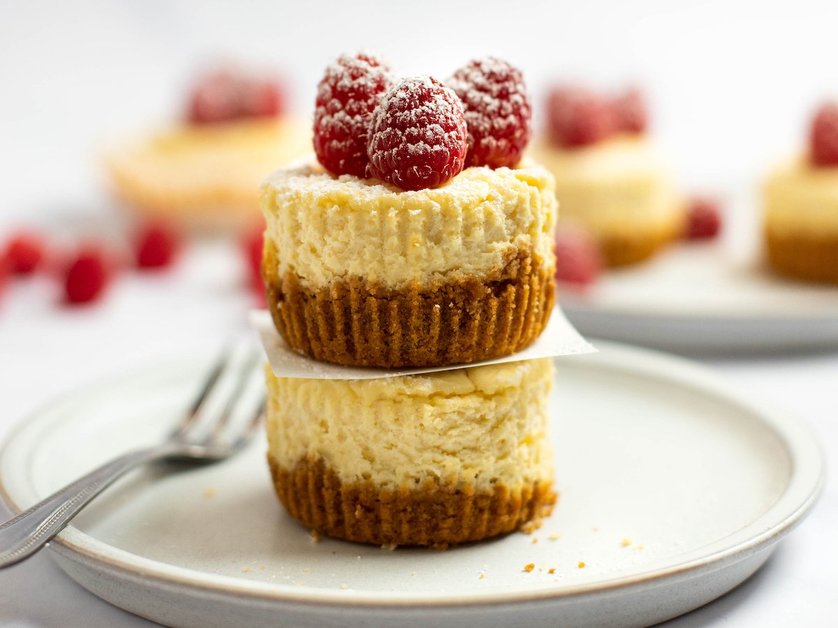 This is a horizontal image looking from the side at a stack of two cupcake-sized cheesecakes. The cheesecakes have lined on them from the muffin liners being pulled away. A small square of white parchment paper is in between the two cheesecakes. On the top cheesecake are three raspberries topped with powdered sugar. The cheesecakes sit on a white plate on a light marble surface. A fork is coming in from the left side of the image and blurred on the side of the plate. More cupcake cheesecakes and raspberries are blurred in the background.