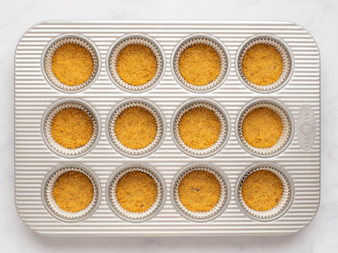 This is an overhead horizontal image of a silver 12 cup muffin pan on a white marble surface. In the pan is a graham cracker cookie crust pressed into the bottom of the muffin liners around them.