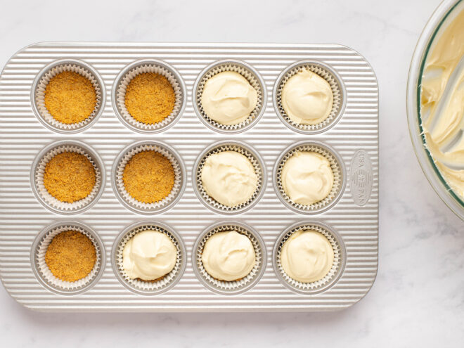 This is an overhead horizontal image of a silver 12 cup muffin pan on a white marble surface. Raw cheesecake batter with muffin liners is in seven of the muffin cups and the remaining cups just have a cookie crust pressed into the bottom. A sliver of a glass bowl with cheesecake better is peeking into the shot from the right side of the image.