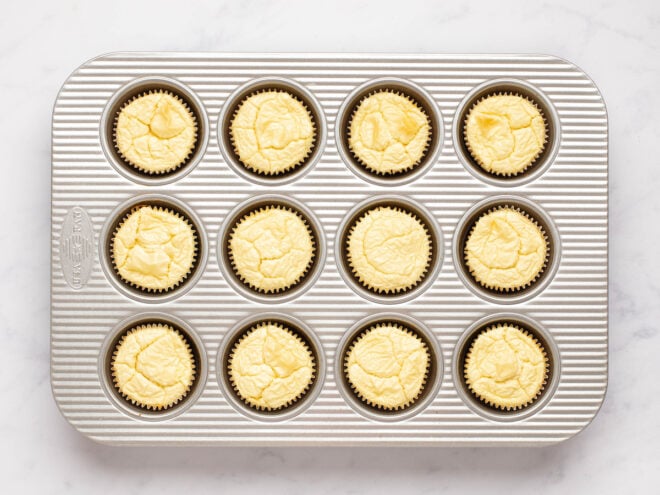This is an overhead horizontal image of a silver 12 cup muffin pan on a white marble surface. In the pan is baked cheesecakes with muffin liners around them.
