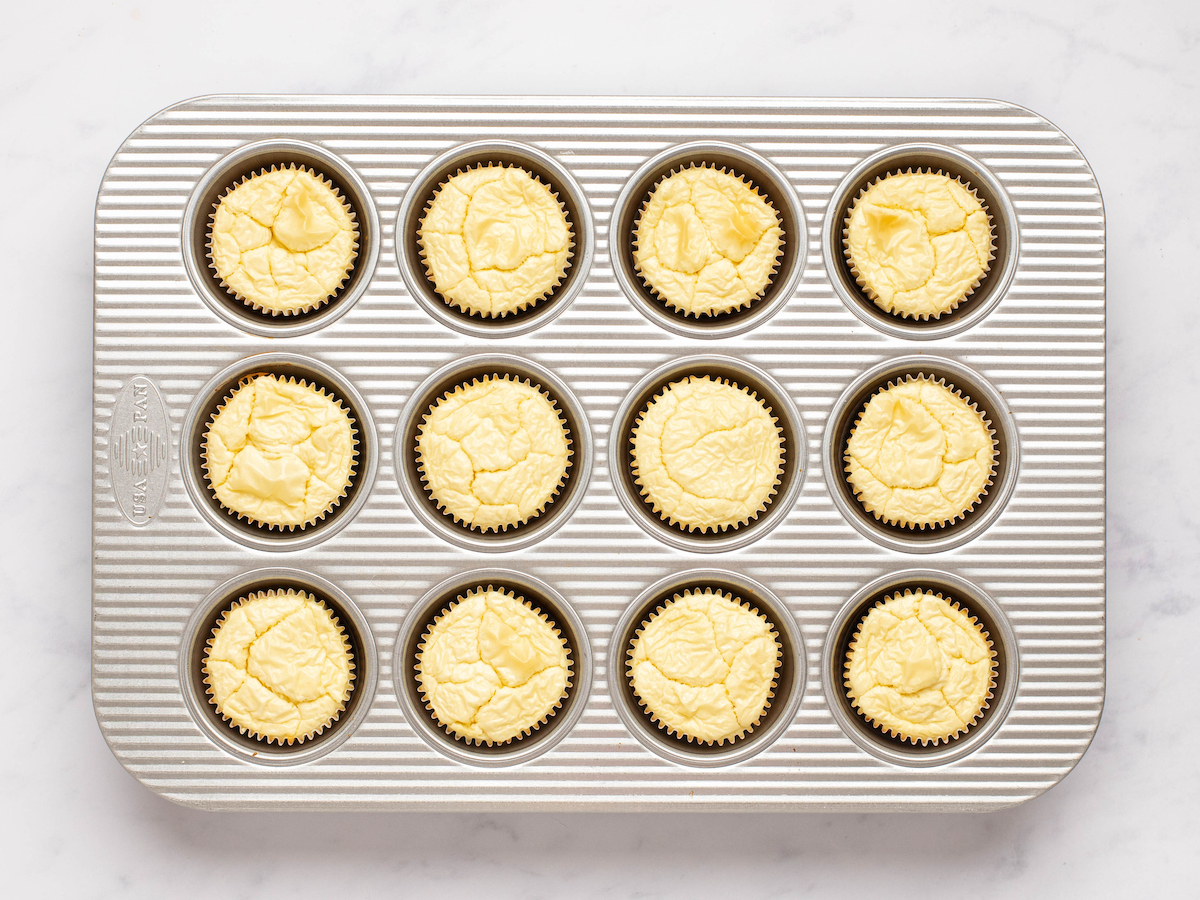 This is an overhead horizontal image of a silver 12 cup muffin pan on a white marble surface. In the pan is baked cheesecakes with muffin liners around them.