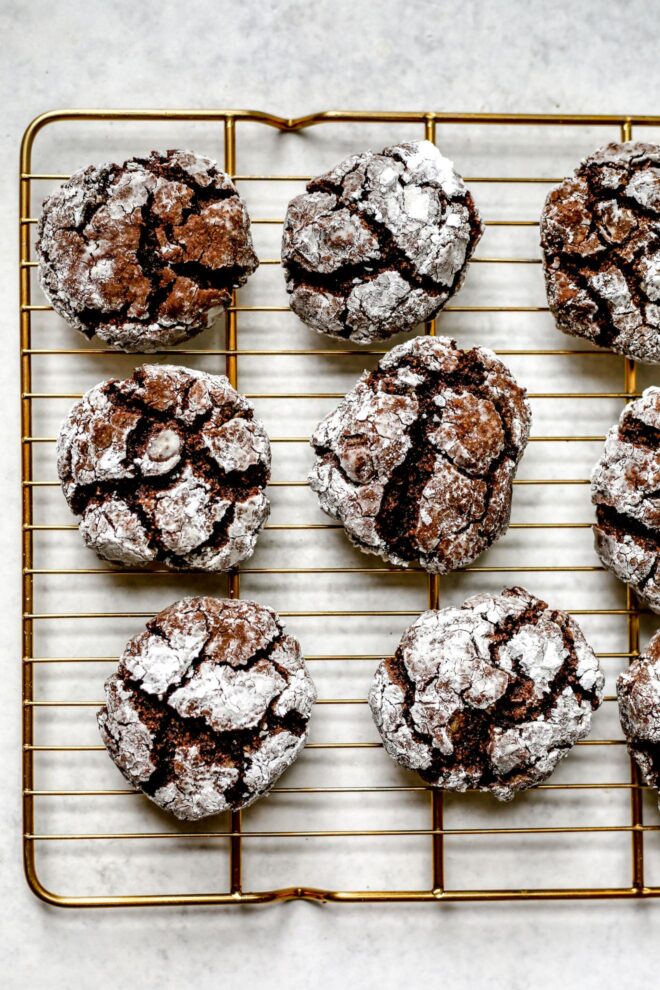 This is an overhead vertical image of nine brownie crinkle cookies coated in powdered sugar on a gold cooling rack. The cooling rack sits on a light grey surface.