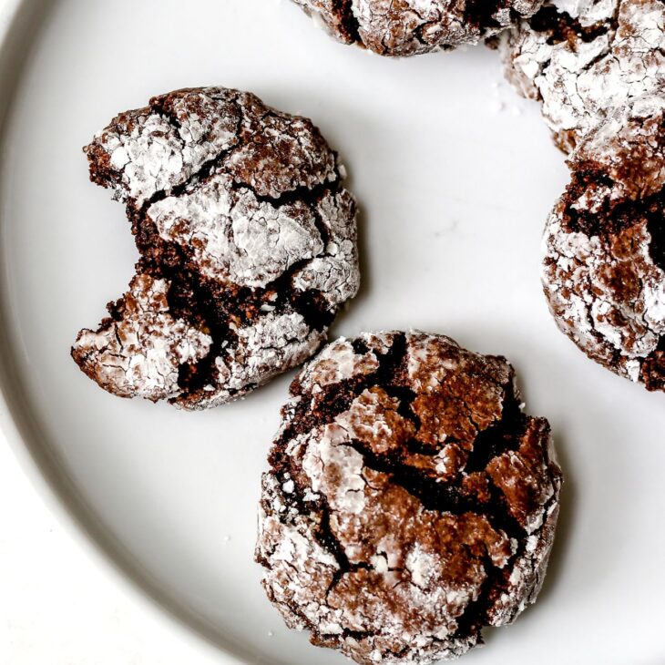 This is a vertical overhead image of a white plate with chocolate brownie crinkle cookies on it. One cookie has a bite taken out. The plate sits on a light grey surface.