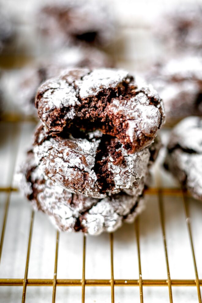 This is a vertical image of a stack of three brownie crinkle cookies on a gold baking sheet. More crinkle cookies are blurred in the background.