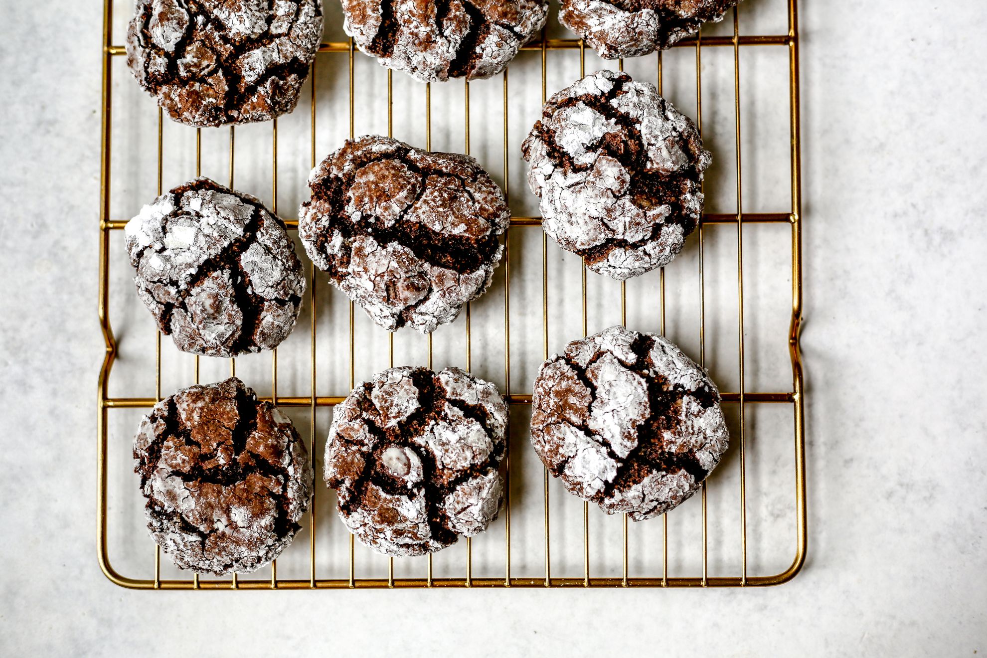 This is an overhead horizontal image of nine brownie crinkle cookies coated in powdered sugar on a gold cooling rack. The cooling rack sits on a light grey surface.