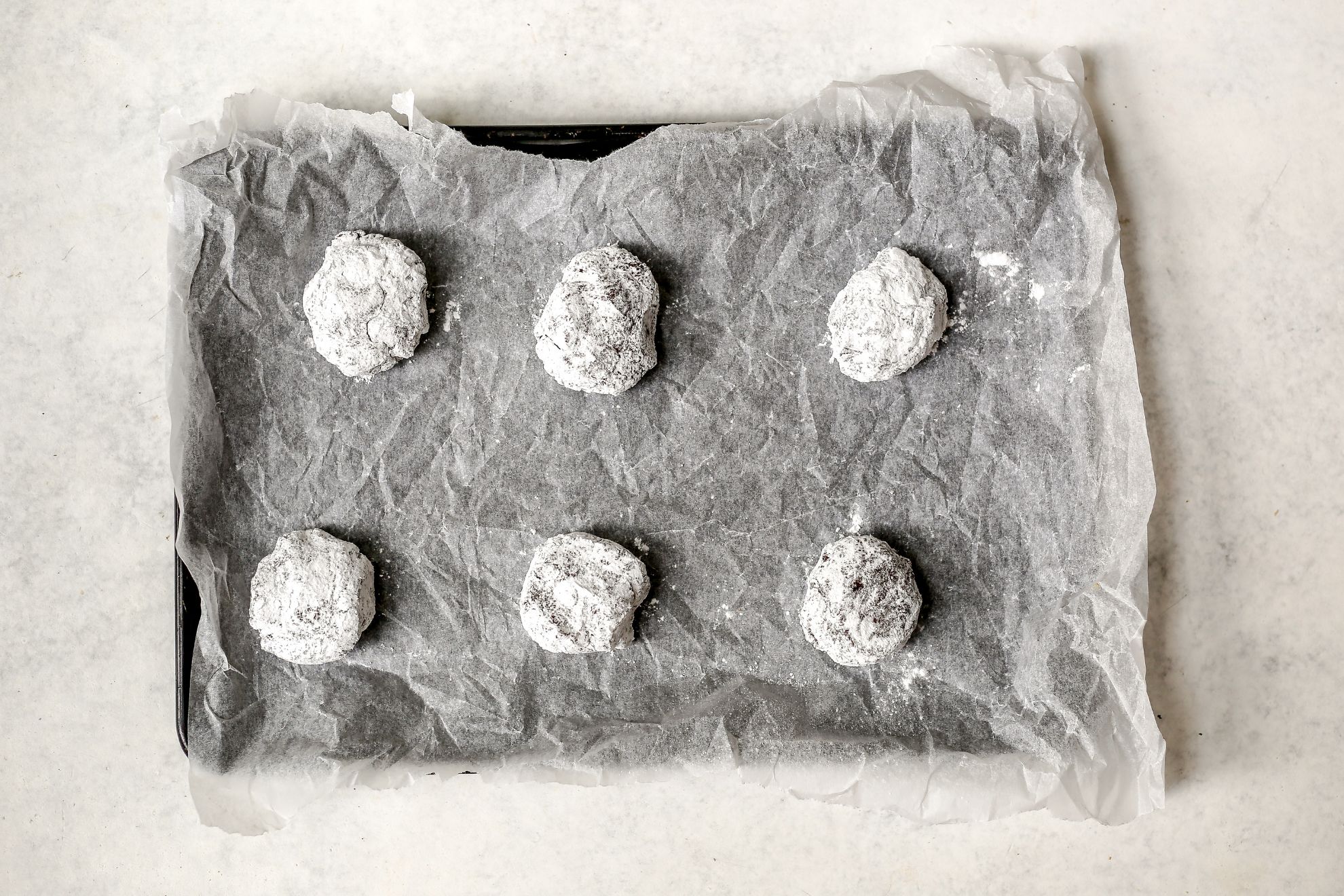 This is an overhead horizontal image of a baking sheet lined with crinkled white parchment paper. Size cookie dough balls coated with powdered sugar are on the baking sheet. The baking sheet sits on a light grey surface.