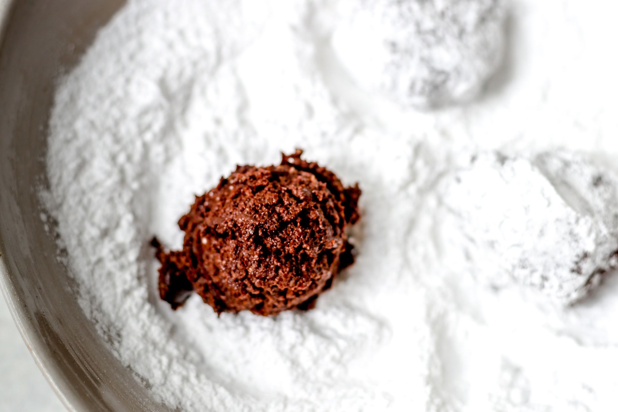This is a horizontal image looking from a side view of powdered sugar in a bowl with balls of chocolate cookie dough in them. Two balls are coated in powdered sugar and the other, closest to the camera is not yet coated, sitting on top of the sugar.