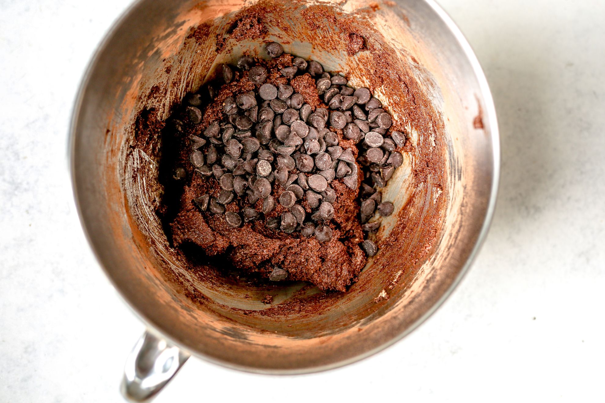 This is an overhead horizontal image of a silver mixing bowl with raw chocolate batter in it. Chocolate chips are on top of the batter. The mixing bowl sits on a light grey/white surface.