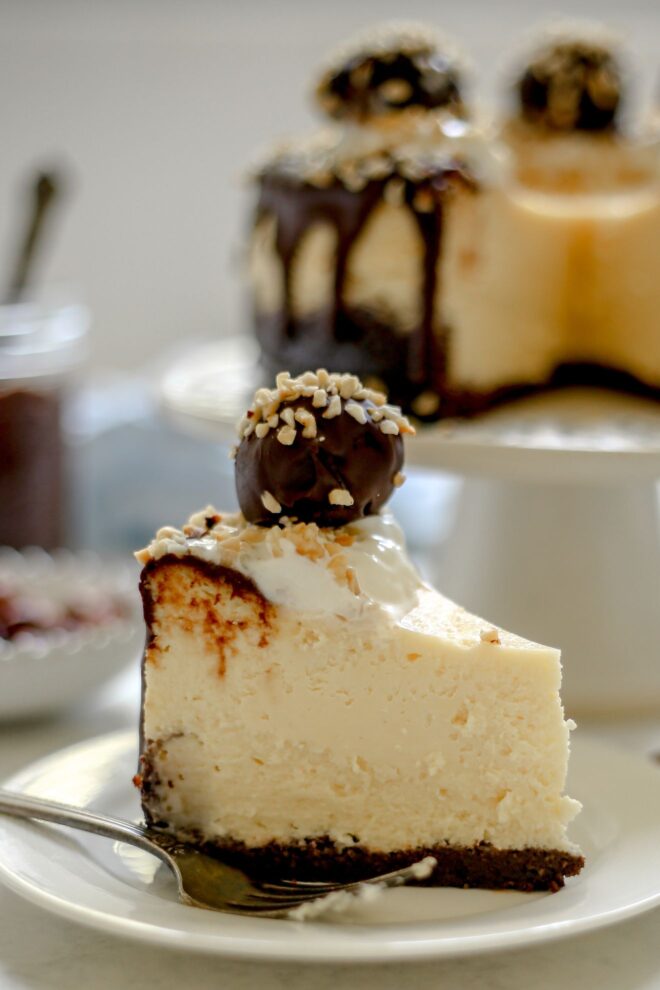 This is a vertical image looking from the side at a slice of cheesecake with a chocolate crust. The slice is topped with chocolate drizzle, whipped cream, chopped nuts and a ferrero rocher candy. The slice is on a white plate on a white surface with a fork leaning on the plate in front of the plate. Blurred in the background is the rest of the cheesecake on a white pedestal and a jar of chocolate with a spoon sticking out of it.