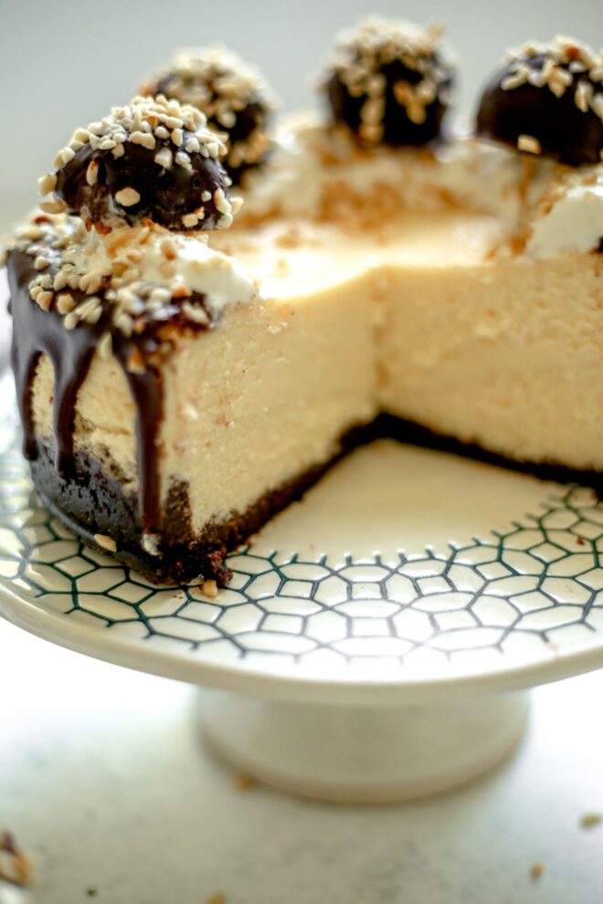 This is a vertical image looking at a cheesecake from the side. The cheesecake is topped with drizzled of chocolate, whipped cream, chopped nuts and ferrero rocher candies. The cheesecake is cut into and the image reveals the center and texture of the cheesecake. The cake sits on a white cake stand with a dark blue.green design around the edges. The cake stand sits on a light grey surface.