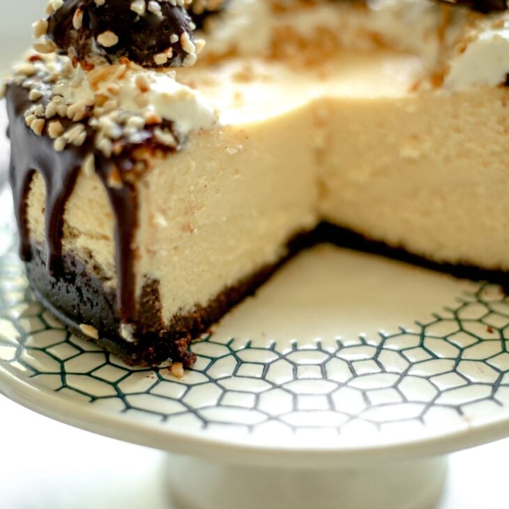 This is a vertical image looking at a cheesecake from the side. The cheesecake is topped with drizzled of chocolate, whipped cream, chopped nuts and ferrero rocher candies. The cheesecake is cut into and the image reveals the center and texture of the cheesecake. The cake sits on a white cake stand with a dark blue.green design around the edges. The cake stand sits on a light grey surface.