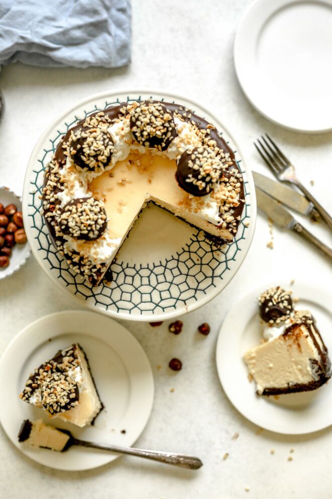 This is an overhead vertical image of a cheesecake topped with whipped cream, chocolate, chopped nuts and ferrero rocher candies. The cheesecake is on a white cake stand with black design detail. Two slices of cheesecake are cut from the cake and on small white plates to the bottom of the image. Beneath the cake stand to the left is a small bowl of hazelnuts and a light blue tea towel to the top left corner. To the right of the cake stand is a couple of plates and forks and knives to the right side of the image.