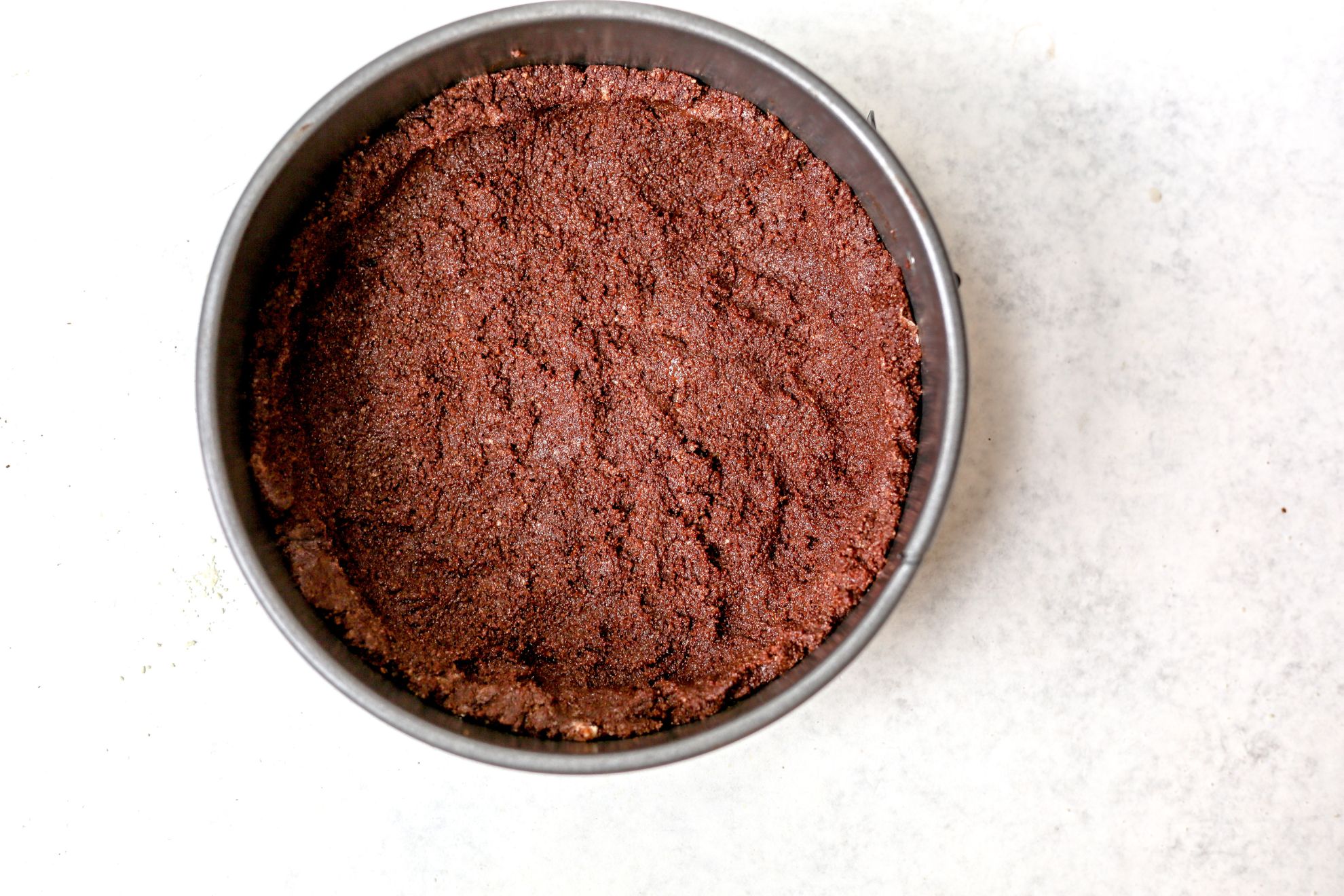 This is an overhead horizontal image of a round springform pan on a light grey surface. In the pan is chocolate cookie crust pressed into the bottom.