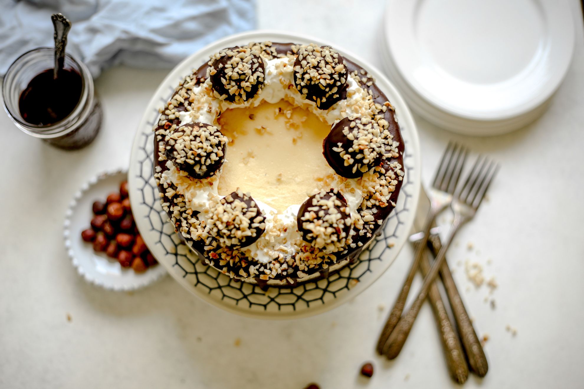 This is an overhead horizontal image of a cheesecake topped with whipped cream, chocolate, chopped nuts and ferrero rocher candies. The cheesecake is on a white cake stand with black design detail. Beneath the cake stand to the left is a small bowl of hazelnuts and a jar of chocolate with a spoon sticking out. To the right of the cake stand is a pile of plates and forks and knives to the right bottom corner of the image.