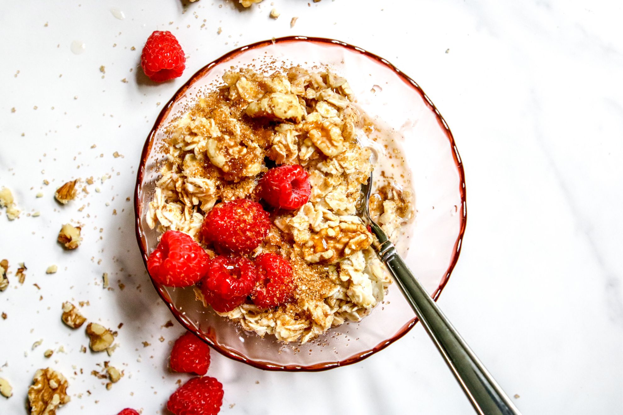 This is an overhead horizontal image of a pink glass bowl with oatmeal in it. A silver spoon is dipping into the oatmeal and leaning against the side of the bowl with the handle pointing to the right bottom corner of the image. The oatmeal is topped with fresh raspberries, walnuts and a sprinkle of brown sugar. The bowl sits on a white marble surface with walnuts and fresh raspberries scattered around it.