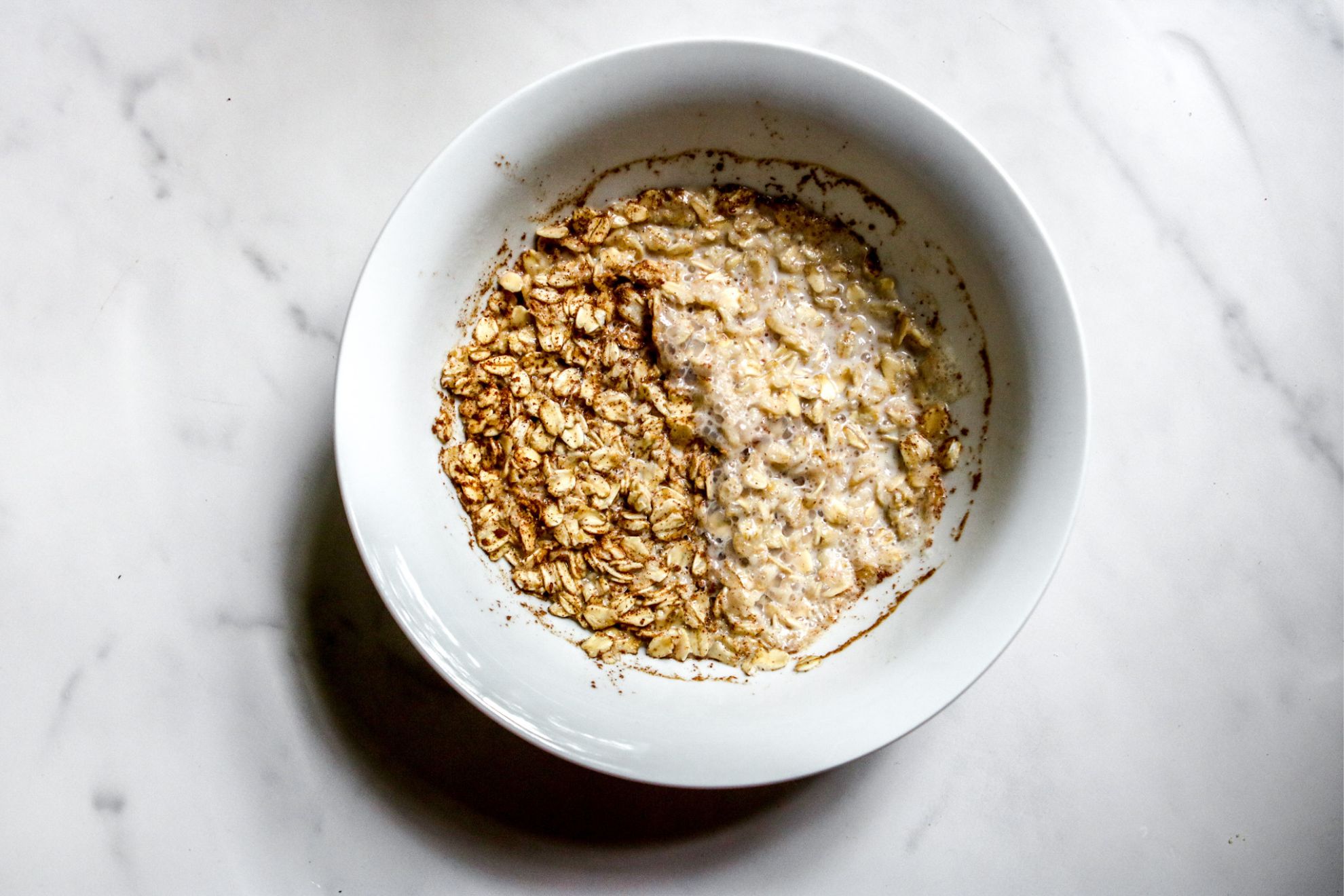 This is an overhead horizontal image of a white bowl on a white marble surface. In the bowl is cooked oatmeal.