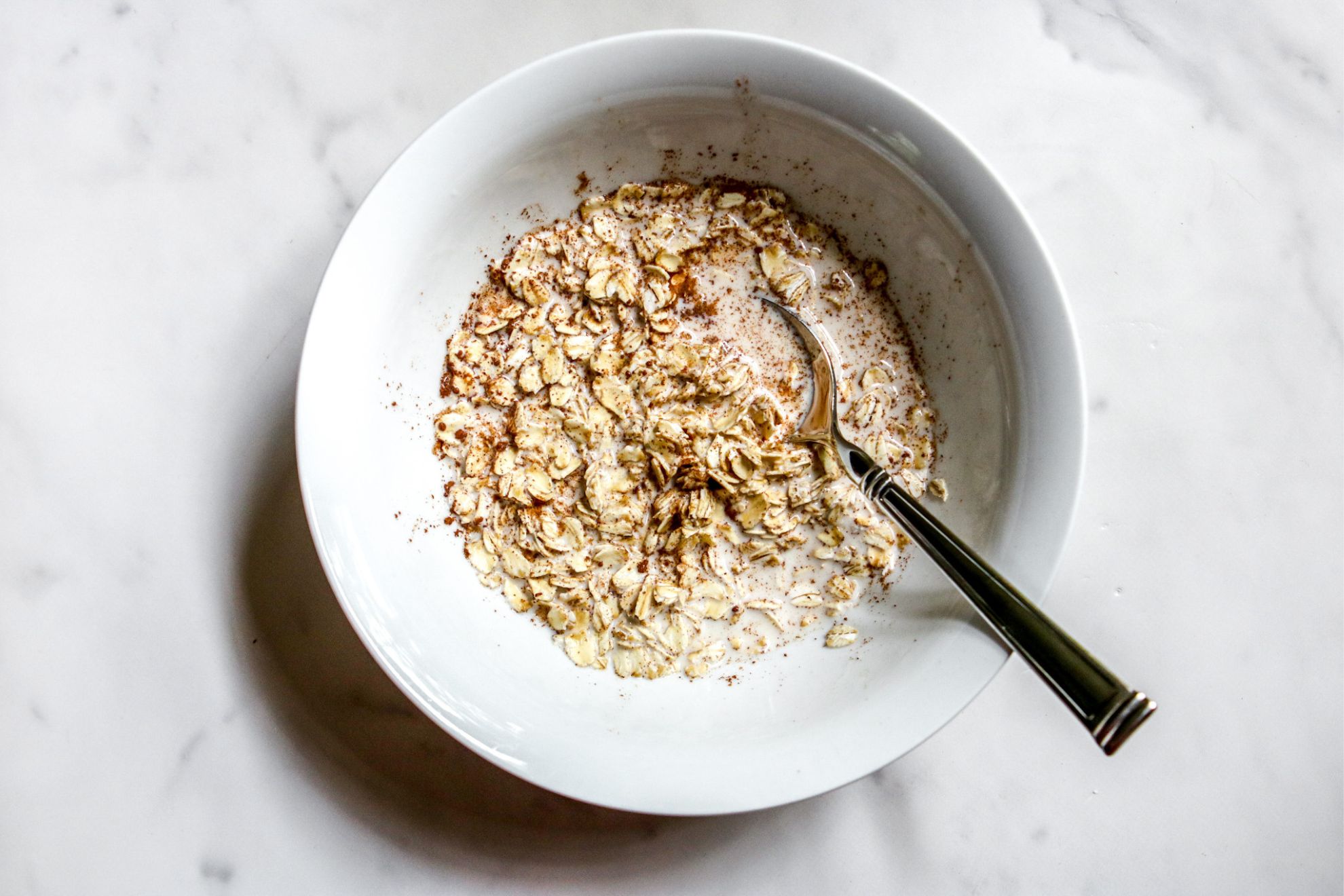 This is an overhead horizontal image of a white bowl on a white marble surface. In the bowl is a milk, spice and oat combination. A silver spoon is in the oatmeal and leaning against the side of the bowl with the handle pointing to the bottom right corner of the image.
