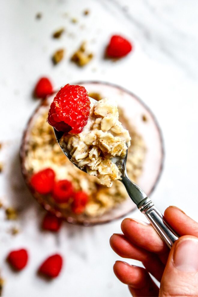 This is a vertical image focusing on a silver spoon with oatmeal and a raspberry on it. A hand is holding the handle of the spoon and is in the bottom right corner of the image. Blurred in the background behind the spoon is a bowl with more oatmeal and raspberries on a white marble surface.