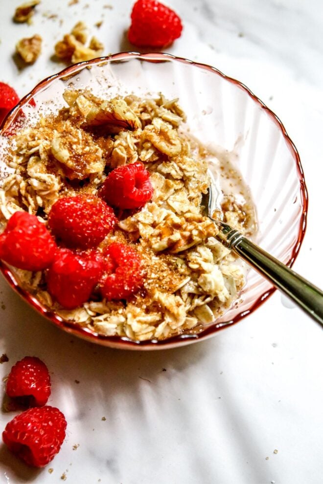 This is a vertical image looking into a pink glass bowl with oatmeal in it. A silver spoon is dipping into the oatmeal and leaning against the side of the bowl with the handle pointing to the right bottom side of the image. The oatmeal is topped with fresh raspberries, walnuts and a sprinkle of brown sugar. The bowl sits on a white marble surface with walnuts and fresh raspberries scattered around it.