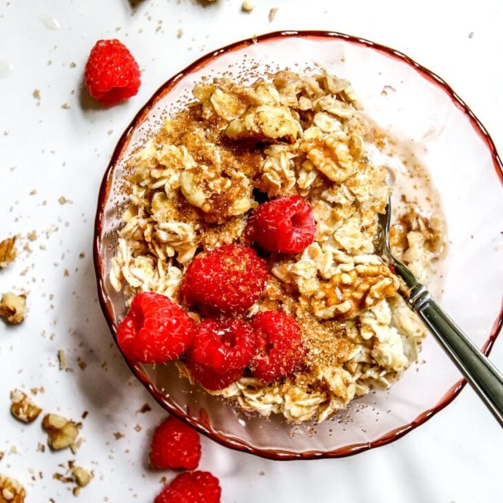 This is an overhead vertical image of a pink glass bowl with oatmeal in it. A silver spoon is dipping into the oatmeal and leaning against the side of the bowl with the handle pointing to the right bottom side of the image. The oatmeal is topped with fresh raspberries, walnuts and a sprinkle of brown sugar. The bowl sits on a white marble surface with walnuts and fresh raspberries scattered around it.