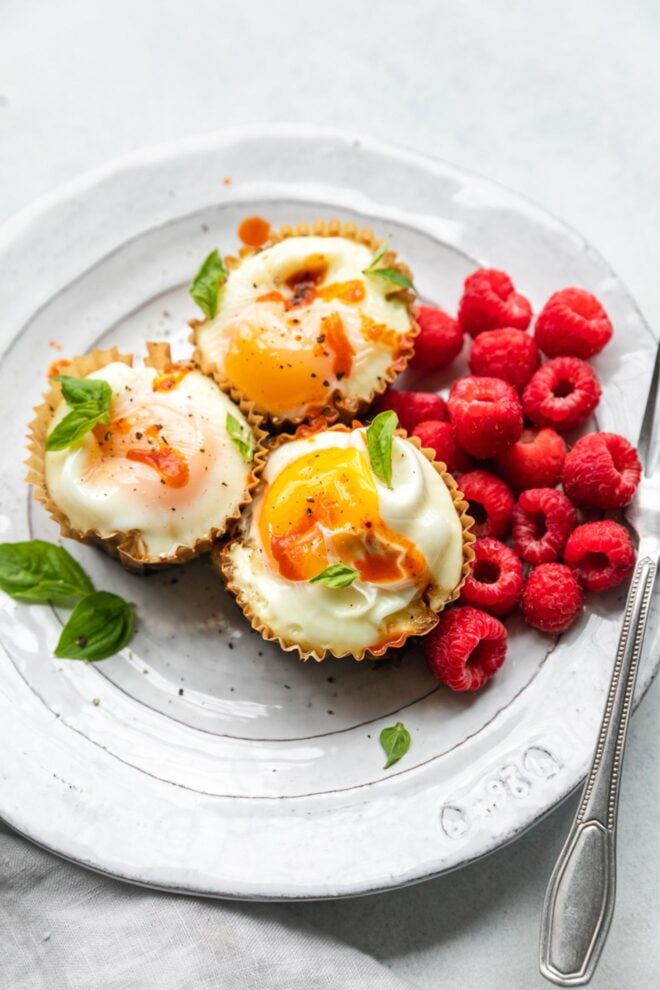 This is a vertical image looking at a 3/4 view onto a white plate with three cooked egg in muffin liners. They are topped with drips of hot sauce and basil leaves on top and around the cups. A couple handfuls of raspberries are to the right of the egg cups. A knife is leaning on the side of the white plate at an angle with the blade cut off on the right side of the image. The plate is on a white surface with a beige cloth to the bottom left corner of the image.