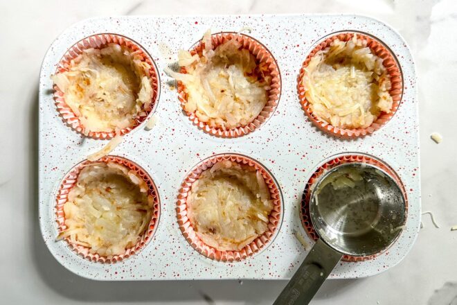 This is an overhead horizontal image of a white six cup muffin tin. The white tin has red speckles on it and is on a white marble surface. In the muffin tin are muffin liners filled halfway with shredded hash browns. A small silver dry measuring cup is sitting in the bottom right muffin tin.