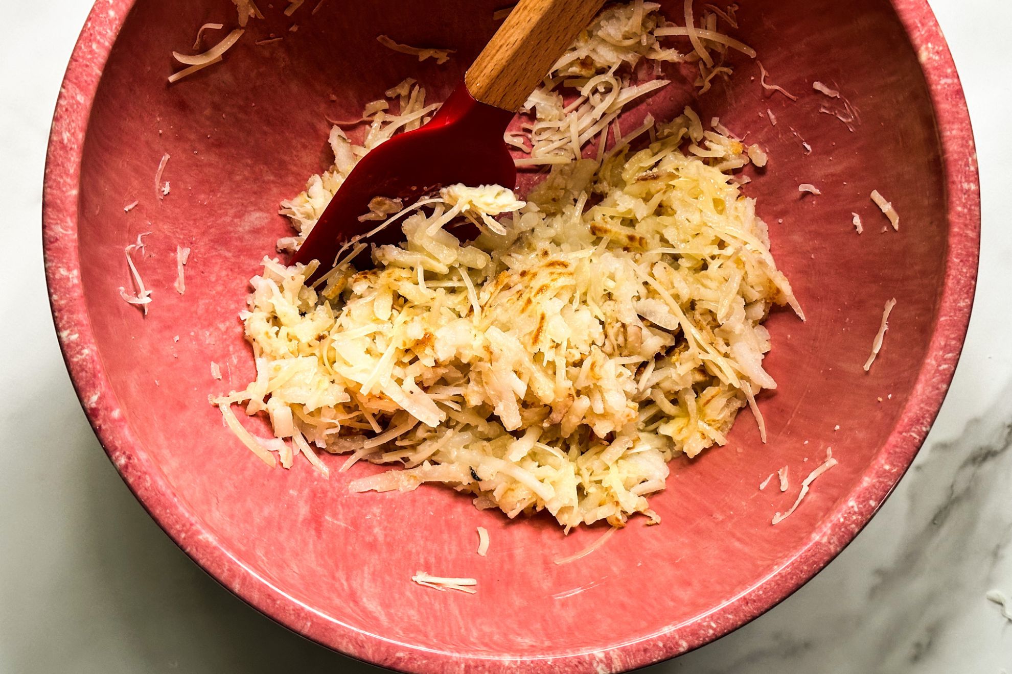 This is an overhead horizontal image of a pink bowl on a white marble surface. In the bowl is a shredded potato mixture with a red rubber spatula with a wooden handle in the bowl and leaning to the top middle of the image.