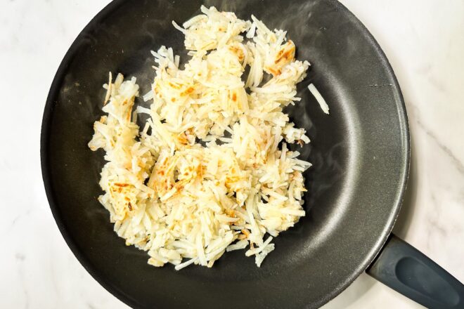 This is an overhead horizontal image looking onto a black skillet with cooked shredded hash browns. Some hash browns are golden brown. The skillet is on a white marble surface.
