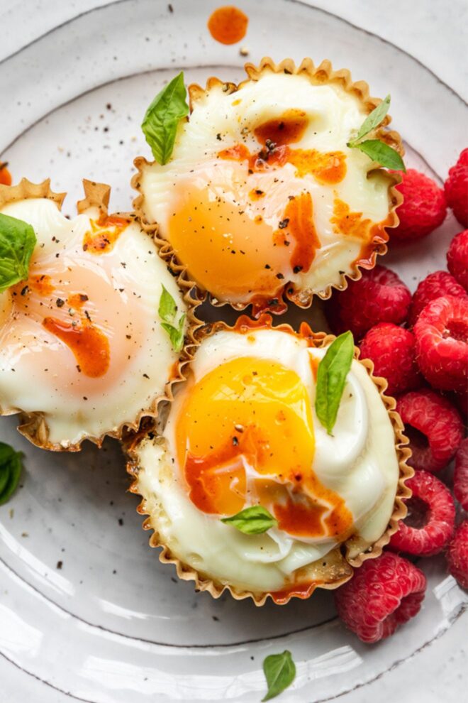 This is a vertical overhead image looking closeup at a white plate with three cooked eggs in muffin liners. They are topped with drips of hot sauce and basil leaves on top and around the cups. A couple handfuls of raspberries are to the right of the egg cups. A knife is leaning on the side of the white plate at an angle with the blade cut off on the right side of the image.