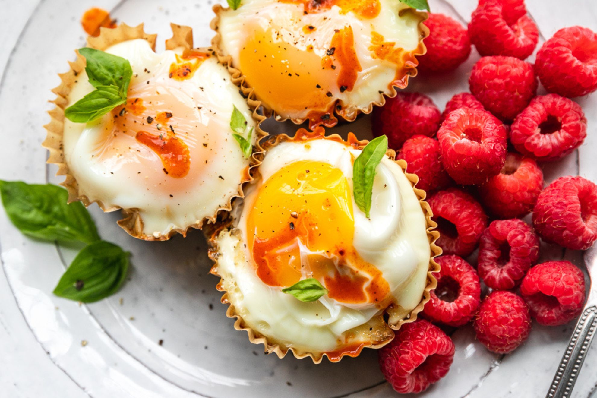 This is a horizontal overhead image looking closeup at a white plate with three egg bites. The egg bites are lined with muffin liners and topped with drips of hot sauce and basil leaves on top and around the cups. A couple handfuls of raspberries are to the right of the egg cups. A knife is leaning on the side of the white plate at an angle with the blade cut off on the right side of the image.