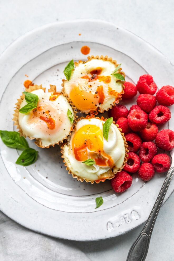 This is an overhead vertical image looking at a white plate with three cooked eggs in a muffin liner. The egg are topped with drips of hot sauce and basil leaves on top and around the cups. A couple handfuls of raspberries are to the right of the egg cups. A knife is leaning on the side of the white plate at an angle with the blade cut off on the right side of the image. The plate is on a white surface with a beige cloth to the bottom left corner of the image.