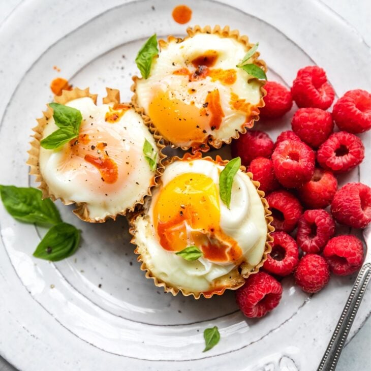 This is an overhead vertical image looking at a white plate with three cooked eggs in muffin liners. The egg bites are topped with drips of hot sauce and basil leaves on top and around the cups. A couple handfuls of raspberries are to the right of the egg cups. A knife is leaning on the side of the white plate at an angle with the blade cut off on the right side of the image. The plate is on a white surface with a beige cloth to the bottom left corner of the image.