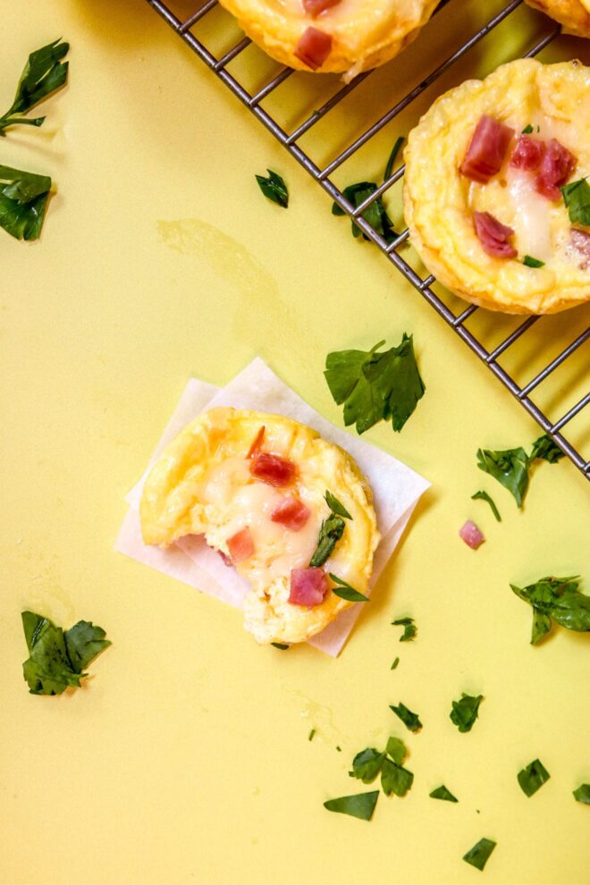 This is an overhead vertical image of a silver cooling rack with muffin sized quiches on it in the top right corner of the image. The quiches are topped with melted cheese, chunks of ham, and chopped parsley. The cooling rack sits on a yellow surface. In the center of the image is a small crustless quiche on a white square of parchment paper. Chopped parsley is sprinkled around the yellow surface.