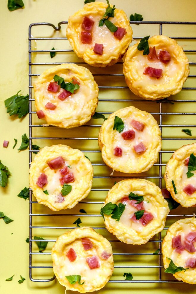 This is an overhead vertical image of a silver cooling rack with muffin sized quiches on it. The quiches are topped with melted cheese, chunks of ham, and chopped parsley. The cooling rack sits on a yellow surface.