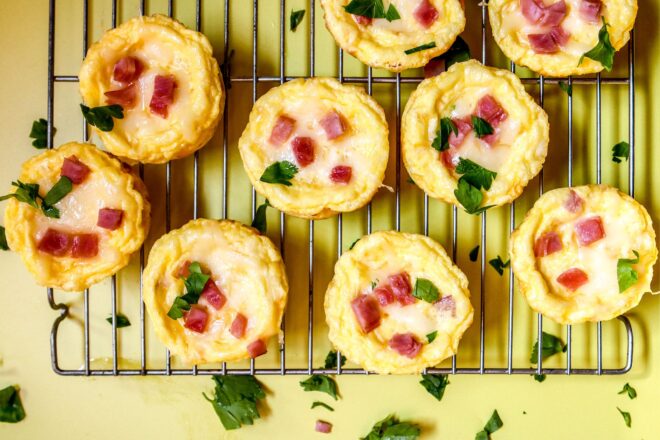 This is an overhead horizontal image of a silver cooling rack with muffin sized quiches on it. The quiches are topped with melted cheese, chunks of ham, and chopped parsley. The cooling rack sits on a yellow surface.