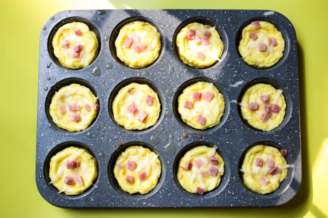 This is an overhead horizontal image of a 12 cup muffin tin. The grey speckled muffin tin has oil sprayed on it and each muffin cup is filled with baked egg quiches topped with chunks of ham and cheese. The muffin tin sits on a yellow surface.