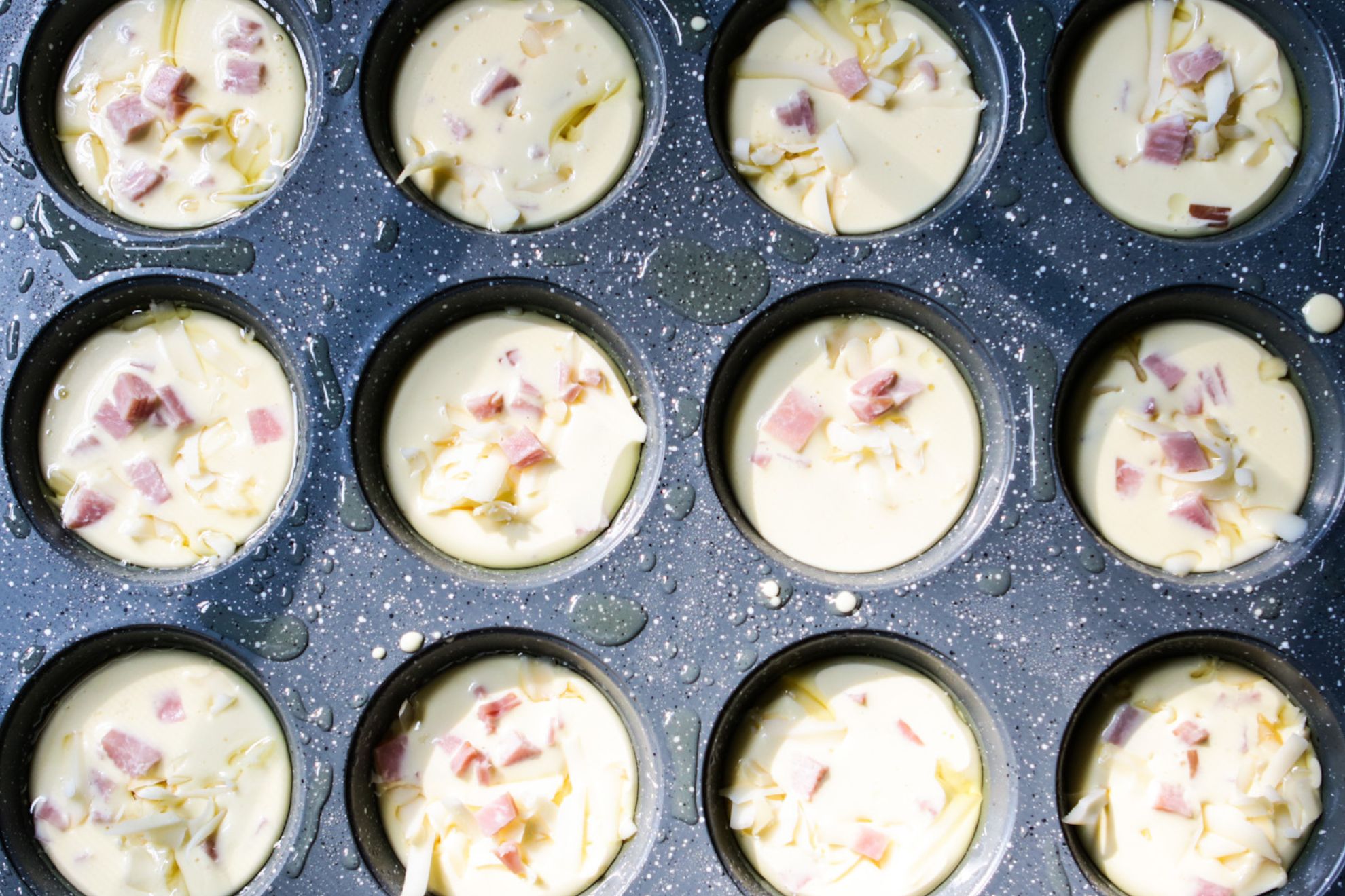 This is an overhead horizontal image of a 12 cup muffin tin. The grey speckled muffin tin has oil sprayed on it and each muffin cup is filled with a raw creamy egg mixture with chunks of ham and shredded cheese.