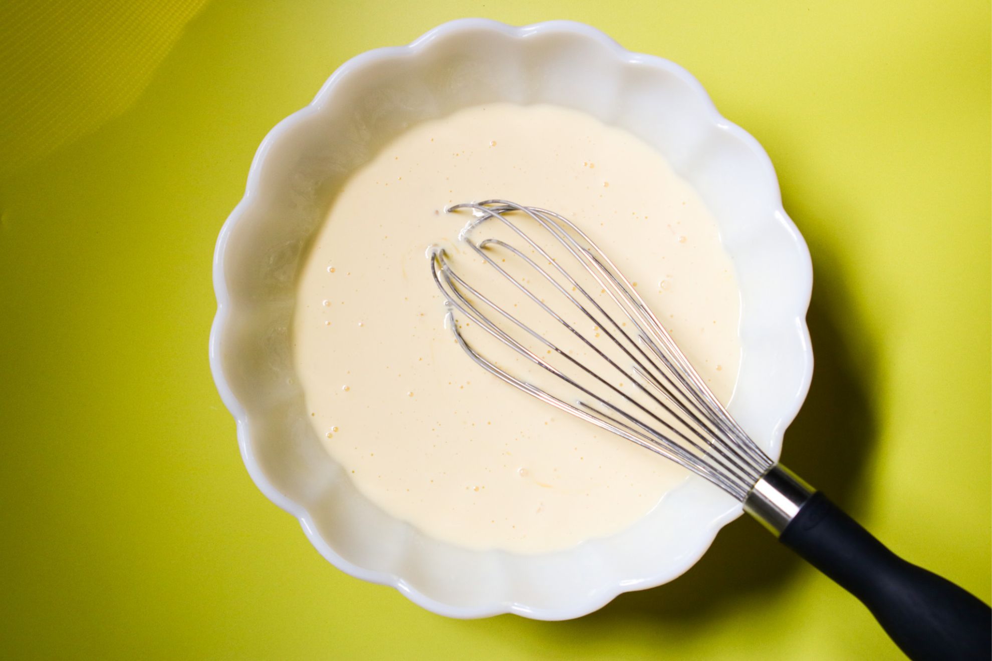 This is an overhead horizontal image of a large scalloped milk glass bowl on a yellow surface. In the bowl is a slightly yellow creamy mixture and a whisk dipped into it. The silver whisk has a black handle and is leaning against the side of the bowl pointing at the bottom right corner of the image.
