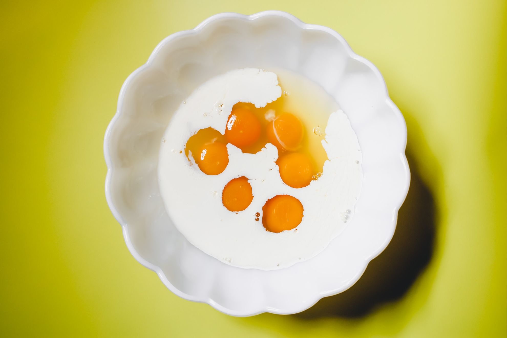 This is an overhead horizontal image of a large scalloped milk glass bowl on a yellow surface. In the bowl are six eggs and heavy cream.