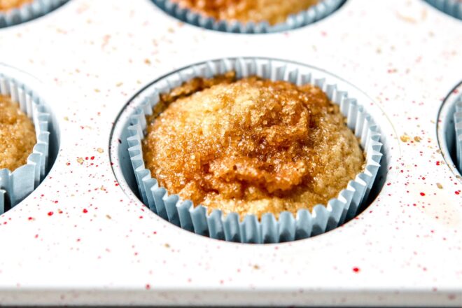 This is a horizontal image looking at a muffin pan from the side. The image focuses on one cavity of the muffin pan. In the muffin cup is a muffin with a sugar topping with a muffin liner around it.