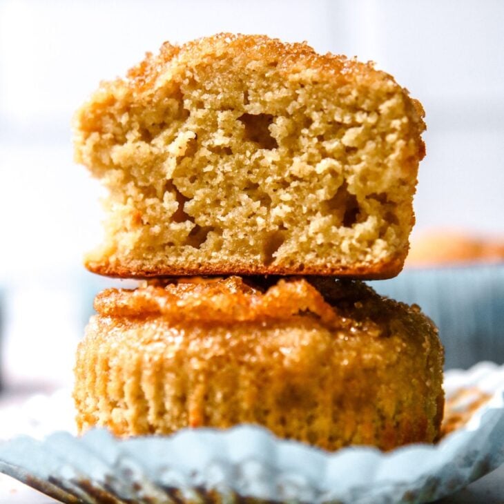 This is a vertical image looking at a stack of two vanilla muffins with a sugary crumb on top. The top muffin is cut in half and both muffins are sitting on a light blue muffin liner. The stack sits on a white surface with a white background and more muffins blurred in the back.