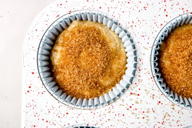 This is an overhead horizontal image looking down onto one cup of a muffin pan. The white pan has red speckles. The muffin cup is lined with a cupcake liner, has raw vanilla batter in it and is topped with brown sugar. The muffin pan sits on a white surface.