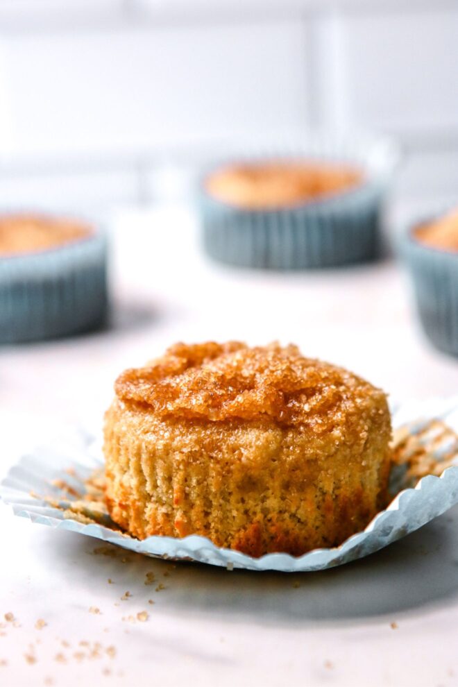This is a vertical image looking at a vanilla muffins with a sugary crumb on top from the side. The muffin is sitting on a light blue muffin liner. The muffins sit on a white surface with a white tile background and more muffins blurred in the back.
