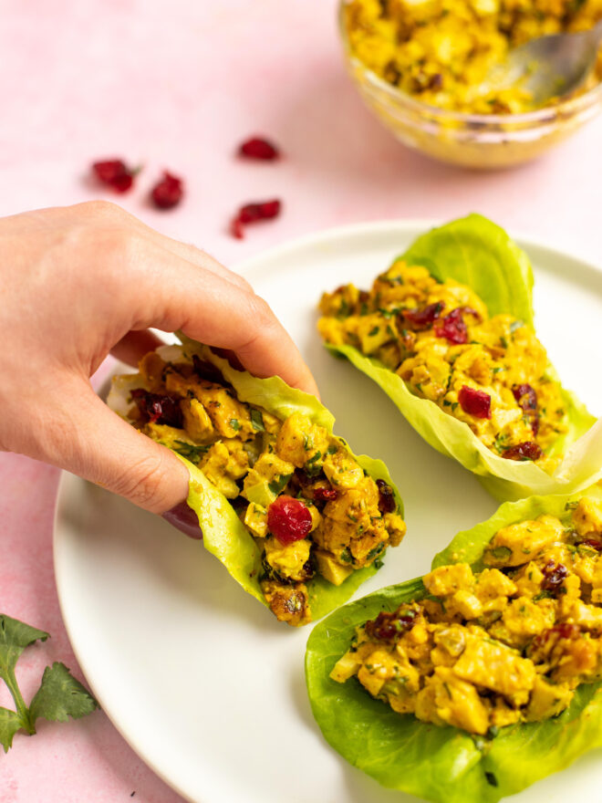 This is a vertical image looking at a white plate from an angled view. On the plate are three lettuce cups filled with a yellow cubed chicken salad with nuts and dried cranberries. A hand is coming from the left side of the image and holding one of the lettuce cups. The plate sits on a light pink surface with a glass bowl of more chicken salad in the background. On the surface behind the plate are a few dried cranberries.