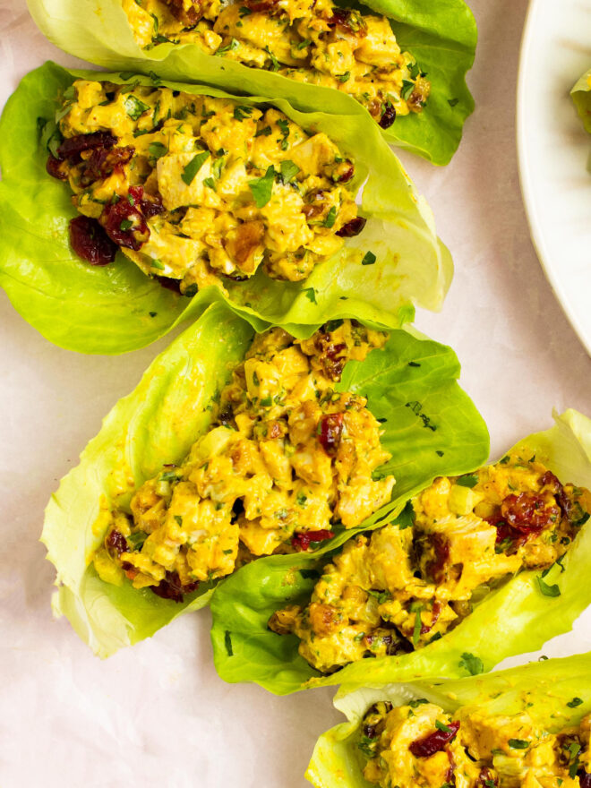 This is a vertical image of lettuce cups on a light pink background. In the lettuce cups is a yellow chicken salad with nuts, dried cranberries and herbs. To the right top corner of the image is a white plate peeking into the image but it's not in the image enough to know what's on the plate.